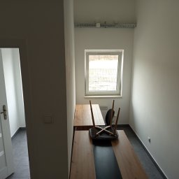 EXPRESS- BUD SPÓŁKA Z OGRANICZONĄ ODPOWIEDZIALNOŚCIĄ - Wnętrze po remoncie: białe ściany, szara podłoga, drewniany blat biurka z czarnym pasem, okno z widokiem na zewnątrz, odwrócone krzesło. Widoczna instalacja elektryczna pod sufitem.