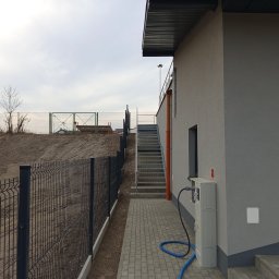 EXPRESS- BUD SPÓŁKA Z OGRANICZONĄ ODPOWIEDZIALNOŚCIĄ - Nowoczesny budynek z szarą elewacją, metalowymi schodami zewnętrznymi i czarnym ogrodzeniem panelowym. Widoczny fragment chodnika z kostki brukowej i instalacja elektryczna.