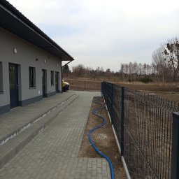 EXPRESS- BUD SPÓŁKA Z OGRANICZONĄ ODPOWIEDZIALNOŚCIĄ - Nowoczesny, grafitowy płot panelowy okalający posesję z budynkiem o szarej elewacji i kostką brukową. W tle widoczne drzewa i pole. Ujęcie z boku.