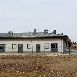 EXPRESS- BUD SPÓŁKA Z OGRANICZONĄ ODPOWIEDZIALNOŚCIĄ - Parterowy budynek z szarą elewacją, ciemnymi oknami i drzwiami, oraz metalowym dachem. Teren wokół budynku jest ogrodzony siatką i częściowo przygotowany pod zagospodarowanie.