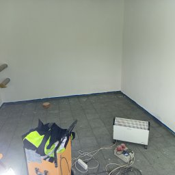 EXPRESS- BUD SPÓŁKA Z OGRANICZONĄ ODPOWIEDZIALNOŚCIĄ - Wnętrze pokoju po malowaniu ścian na biało, z widocznymi śladami farby na szarych płytkach podłogowych i niebieską taśmą malarską przy podłodze. Grzejnik elektryczny i narzędzia.