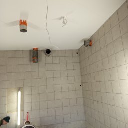 EXPRESS- BUD SPÓŁKA Z OGRANICZONĄ ODPOWIEDZIALNOŚCIĄ - Wnętrze w trakcie remontu: szare płytki na ścianach, widoczne rury wentylacyjne zabezpieczone taśmą, przewody elektryczne i fragment osoby w dolnym rogu kadru.