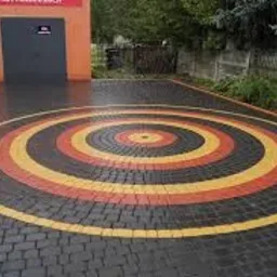 Kolorowa nawierzchnia z kostki brukowej ułożona w koncentryczne okręgi: żółty, pomarańczowy i szary, na tle budynku z ciemnoszarą bramą garażową i zielenią drzew.