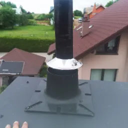 Montaż komina systemowego na dachu, widoczne elementy mocujące i uszczelniające, w tle budynki mieszkalne i zieleń.