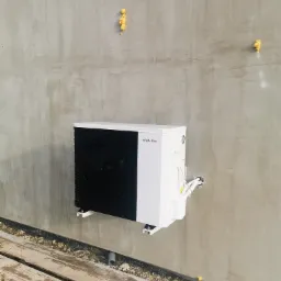 Biała pompa ciepła marki Hewalex zamontowana na szarej, surowej ścianie z widocznymi śladami pianki montażowej, z kablami wychodzącymi ze ściany i fragmentem drewnianego podłoża w dolnej części kadru.