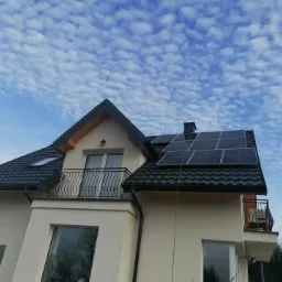 Dom jednorodzinny z zamontowanymi panelami słonecznymi na dachu pokrytym ciemną dachówką, widok z dołu na tle błękitnego nieba z chmurami.