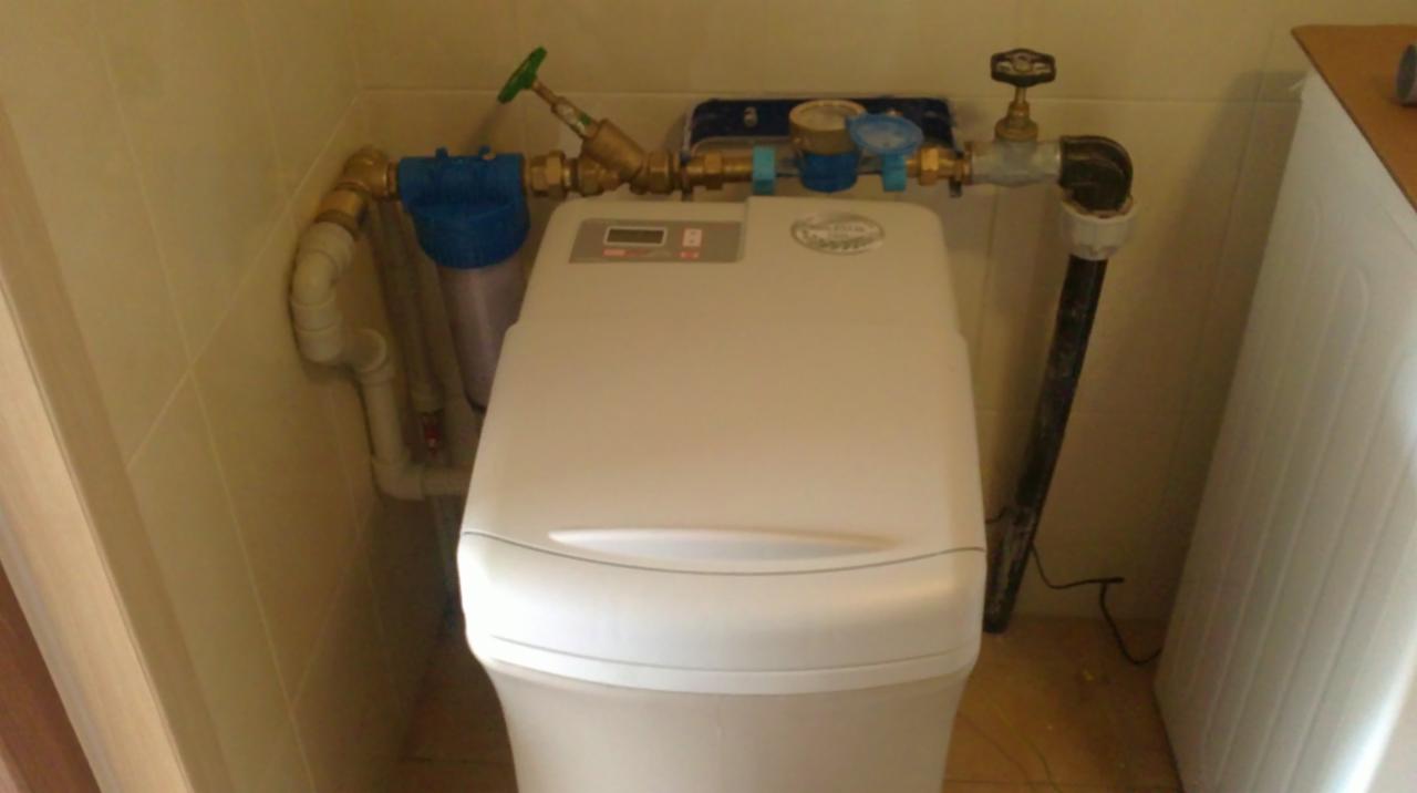 Biały zmiękczacz wody zainstalowany w łazience, widoczne rury, filtry i zawory. Całość otoczona beżowymi płytkami ceramicznymi.