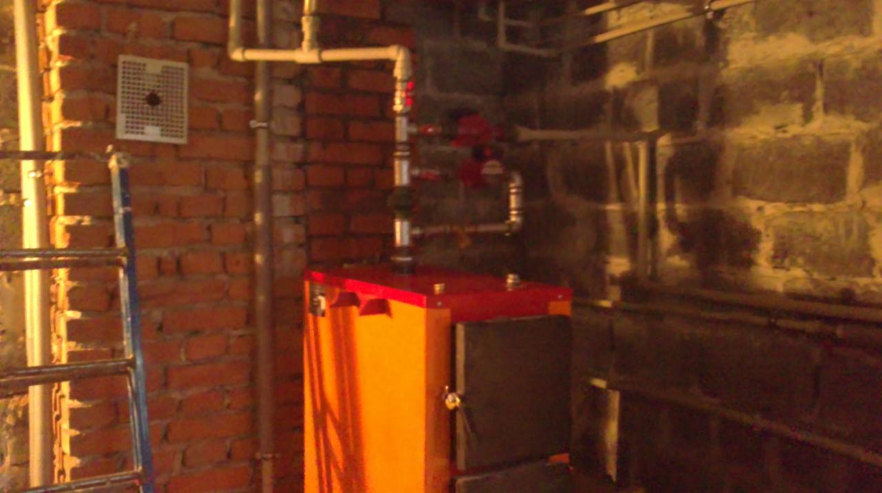 Pomarańczowy piec CO z czerwoną górną częścią, rurami i zaworami na tle surowej ceglanej ściany i drabiny, w pomieszczeniu o słabym oświetleniu.