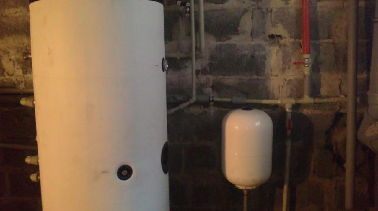 Biały zasobnik ciepłej wody użytkowej i naczynie przeponowe w piwnicy z instalacją hydrauliczną na tle ściany z cegły. Widoczne rury i zawory.
