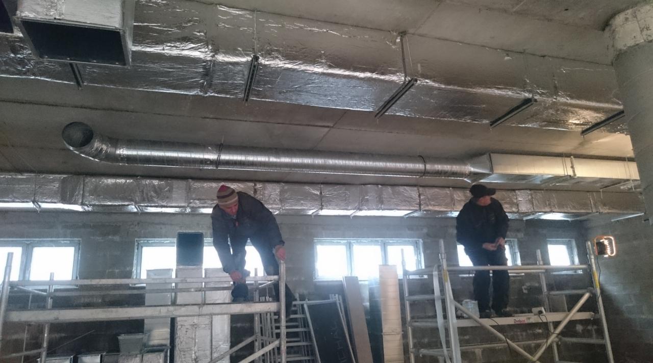Dwóch monterów na rusztowaniach instaluje srebrne rury wentylacyjne pod sufitem w surowym pomieszczeniu z oknami.