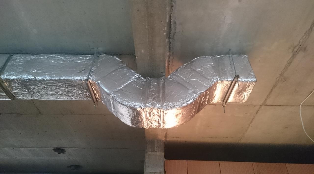Izolowany przewód wentylacyjny w kształcie litery U, zamocowany do sufitu. Srebrna folia izolacyjna, betonowy sufit i fragment ściany z cegieł.