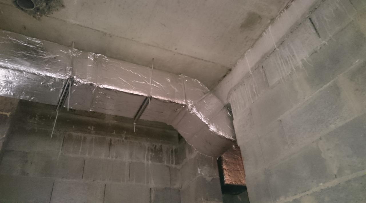 Instalacja wentylacyjna z izolacją, poprowadzona pod sufitem w surowym pomieszczeniu z betonowych bloczków. Widoczne elementy mocujące i izolacja srebrną folią.