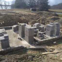 Wznoszenie fundamentów z bloczków betonowych w Olecku, widoczne zbrojenie i przygotowany teren pod budowę, w tle jezioro.