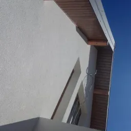 Fragment elewacji budynku z widoczną strukturą tynku, drewnianym podbitką dachu i metalową rynną, uchwycony pod kątem w słoneczny dzień.