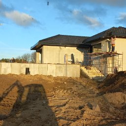 KASBUD Prace Brukarskie Kacper Jaszczerzewski - Budowa domu jednorodzinnego z ciemną dachówką, widoczne fundamenty, ściany w stanie surowym, rusztowania i pracownik na nich. W tle błękitne niebo z chmurami.