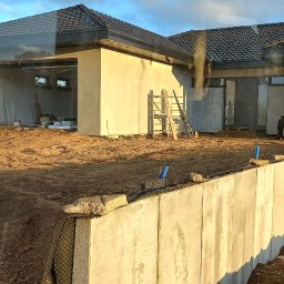 KASBUD Prace Brukarskie Kacper Jaszczerzewski - Dom w trakcie budowy z widocznymi fundamentami i ścianami w stanie surowym. Na pierwszym planie betonowy mur oporowy, w tle rusztowania i drabiny.