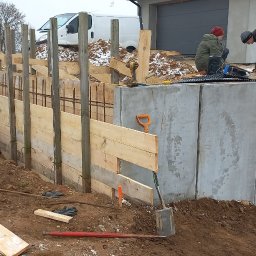 KASBUD Prace Brukarskie Kacper Jaszczerzewski - Wznoszenie fundamentów: pracownicy montują szalunek z desek i zbrojenie. Widoczny betonowy mur, narzędzia i częściowo ośnieżone otoczenie budowy.
