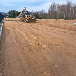 KASBUD Prace Brukarskie Kacper Jaszczerzewski - Żółta koparko-ładowarka wyrównuje teren pod budowę, widoczne ślady opon na ziemi. W tle drzewa i ogrodzenie panelowe.