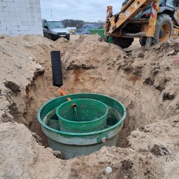 KASBUD Prace Brukarskie Kacper Jaszczerzewski - Montaż zielonego zbiornika w wykopie ziemnym, prawdopodobnie elementu przydomowej oczyszczalni ścieków. W tle koparka i budynek w trakcie budowy.