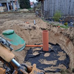 KASBUD Prace Brukarskie Kacper Jaszczerzewski - Montaż przydomowej oczyszczalni ścieków: wykop z rurą drenarską, zbiornik z zieloną pokrywą i czarna geowłóknina. W tle budynek w trakcie budowy.