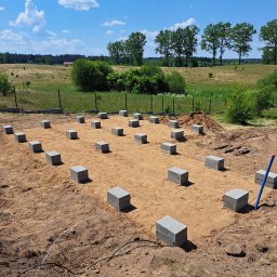 KASBUD Prace Brukarskie Kacper Jaszczerzewski - Przygotowany teren pod fundamenty z ustawionymi bloczkami betonowymi. Widok na pole i drzewa w tle, błękitne niebo z chmurami.