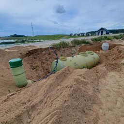KASBUD Prace Brukarskie Kacper Jaszczerzewski - Montaż zbiornika na szambo betonowe w wykopie, widoczne elementy instalacji i krajobraz z domami w tle, pochmurne niebo.