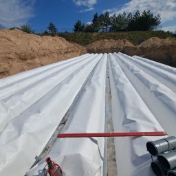 KASBUD Prace Brukarskie Kacper Jaszczerzewski - Drenaż rozsączający: rzędy rur owiniętych białą geowłókniną w wykopie, poziomica i rury PCV obok. Błękitne niebo i drzewa w tle. Elementy instalacji sanitarnej.