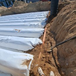 KASBUD Prace Brukarskie Kacper Jaszczerzewski - Instalacja drenażu rozsączającego z rur owiniętych geowłókniną w wykopie, widok z góry z perspektywy osoby stojącej w wykopie, z widoczną łopatą i fragmentem budynku w tle.