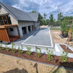 KASBUD Prace Brukarskie Kacper Jaszczerzewski - Nowoczesny dom z szarej kostki brukowej ułożonej w jodełkę, obrzeża z jasnej kostki. Rabaty z korą i zielenią. W tle budynek z garażami i fragment ogrodu z mini koparką.