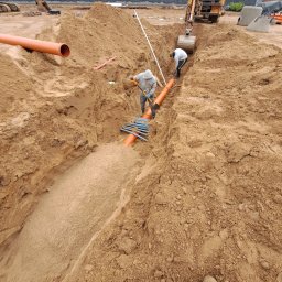 KASBUD Prace Brukarskie Kacper Jaszczerzewski - Montaż pomarańczowych rur w wykopie przez dwóch pracowników, z koparką w tle. Widoczne elementy złączek i narzędzia. Prace ziemne w toku, teren budowy.