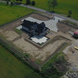 KASBUD Prace Brukarskie Kacper Jaszczerzewski - Widok z lotu ptaka na dom z czarnym dachem i jasną elewacją, otoczony terenem w trakcie zagospodarowania, z widoczną kostką brukową i ogrodzeniem.