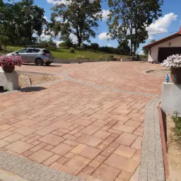 Ułożona z czerwonej kostki brukowej, zakrzywiona ścieżka prowadząca do domu z garażem, z elementami dekoracyjnymi z drobnego żwiru, w tle zaparkowany samochód i trawnik.