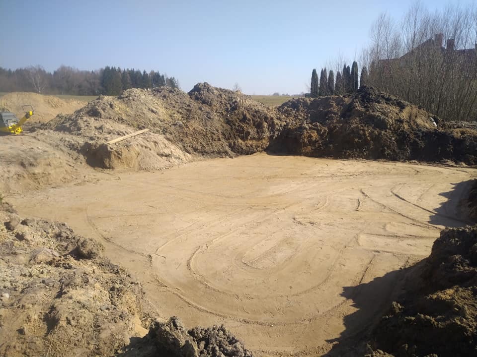 Wykonany wykop pod fundamenty z widocznymi śladami po zagęszczarce gruntu, otoczony usypanymi hałdami ziemi i zielenią w tle.