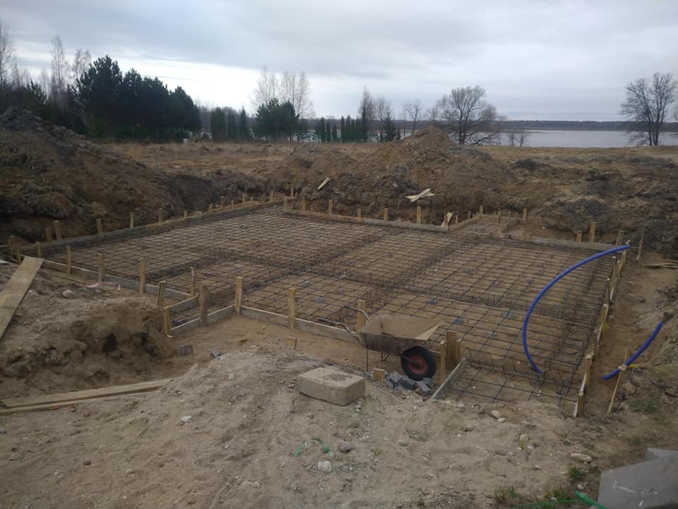 Zbrojenie fundamentów w wykopie, widoczne szalunki drewniane, siatka zbrojeniowa, taczka i niebieski przewód, w tle jezioro i drzewa.