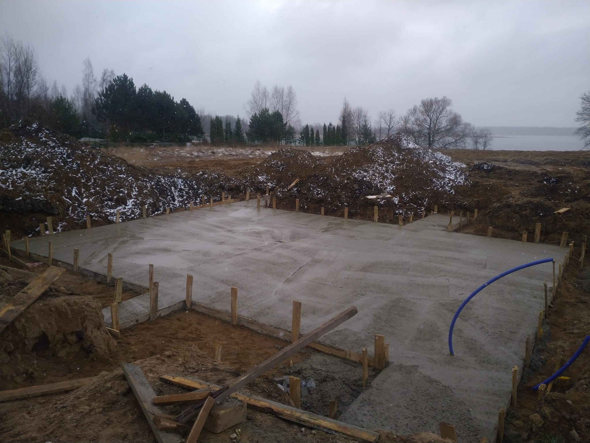 Wylana płyta fundamentowa z drewnianym szalunkiem, widoczne zwały ziemi z resztkami śniegu w tle i fragment jeziora w oddali, pochmurny dzień.
