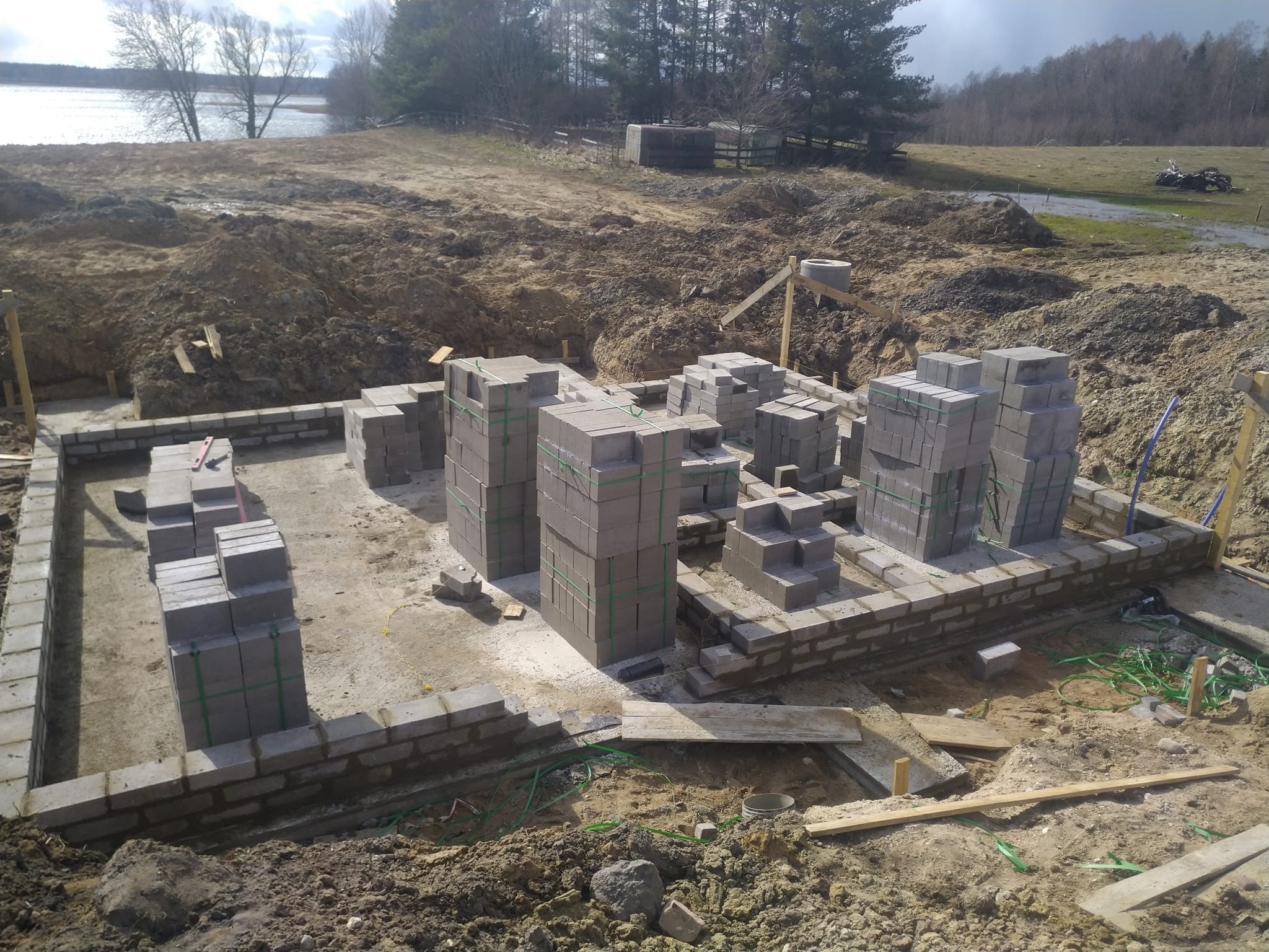 Wznoszenie fundamentów z bloczków betonowych w Olecku, widoczne zbrojenie i przygotowany teren pod budowę, w tle jezioro.