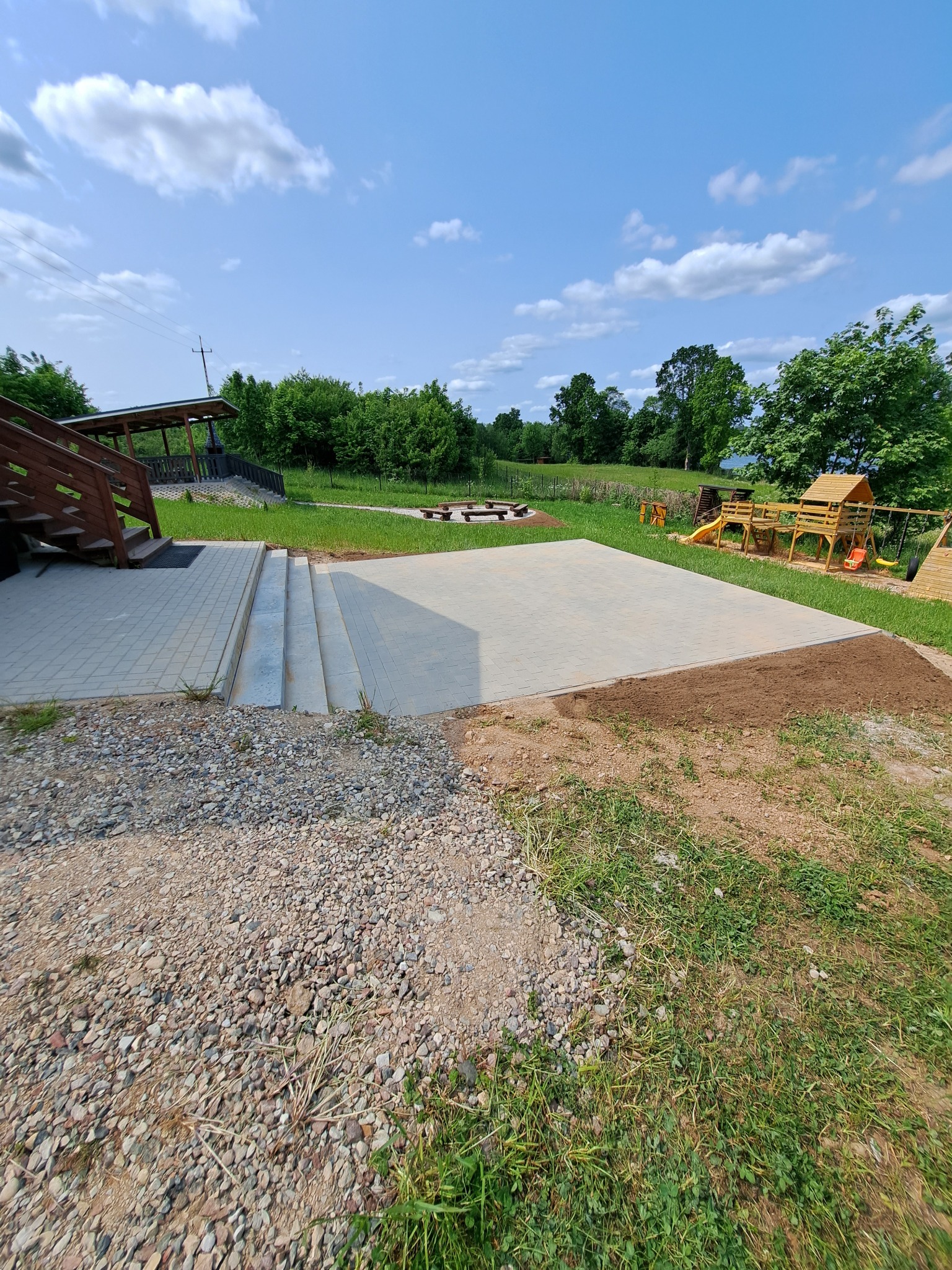 Jasnoszara kostka brukowa ułożona na placu zabaw, z widocznymi schodami i elementami drewnianej konstrukcji. Tło to zieleń trawy i drzew pod błękitnym niebem.