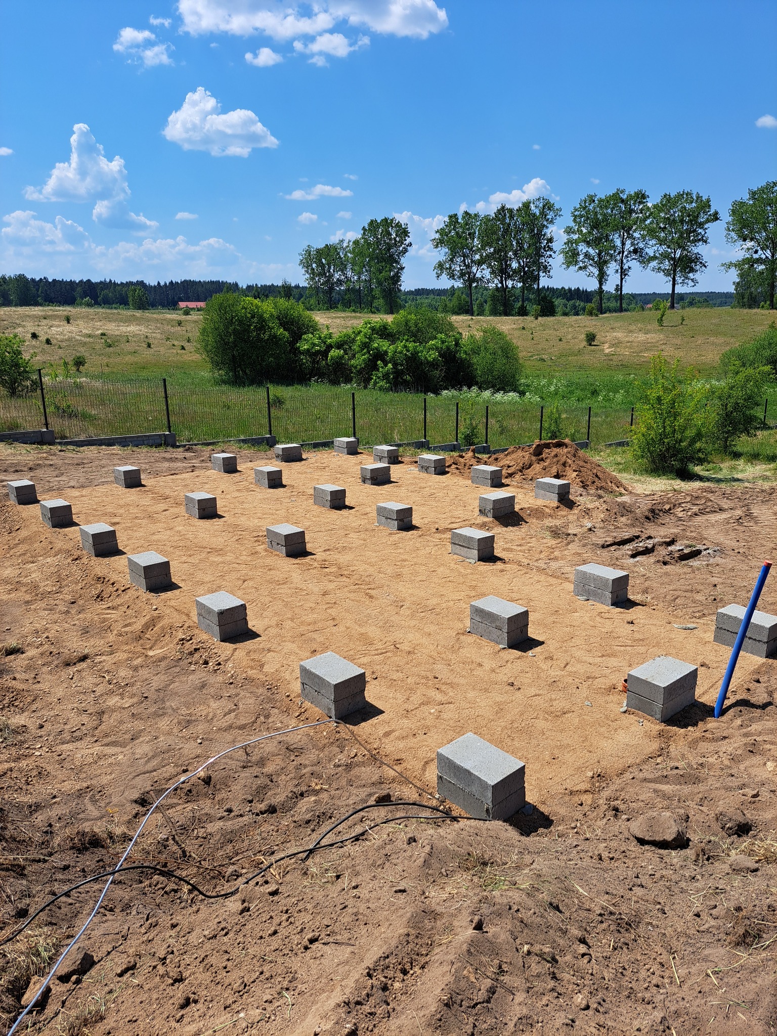Przygotowany teren pod fundamenty z ustawionymi bloczkami betonowymi. Widok na pole i drzewa w tle, błękitne niebo z chmurami.
