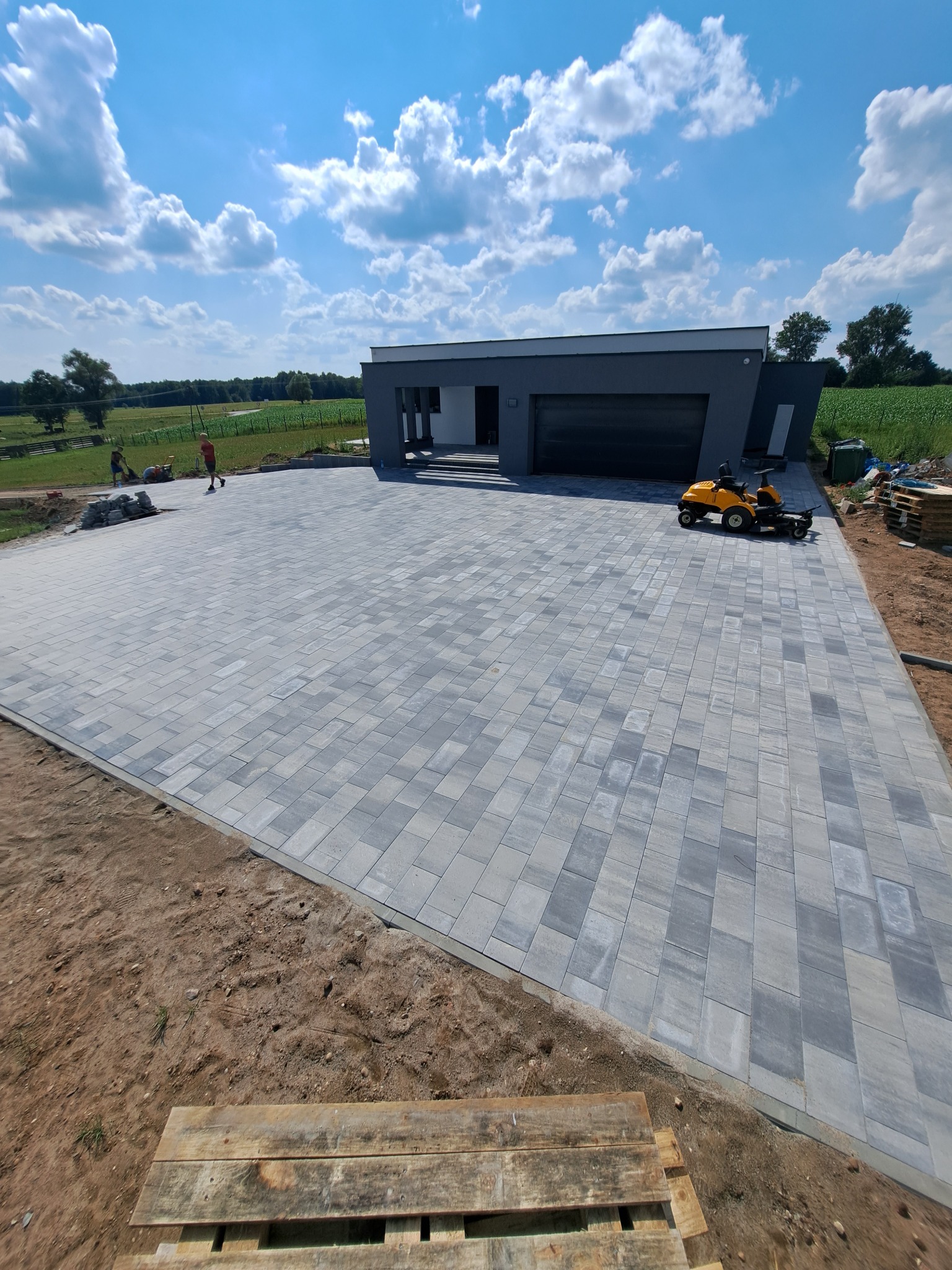 Nowoczesny podjazd z szarej kostki brukowej ułożonej we wzór prostokątów, z domem w tle i kosiarką samojezdną. Widoczni pracownicy w oddali.