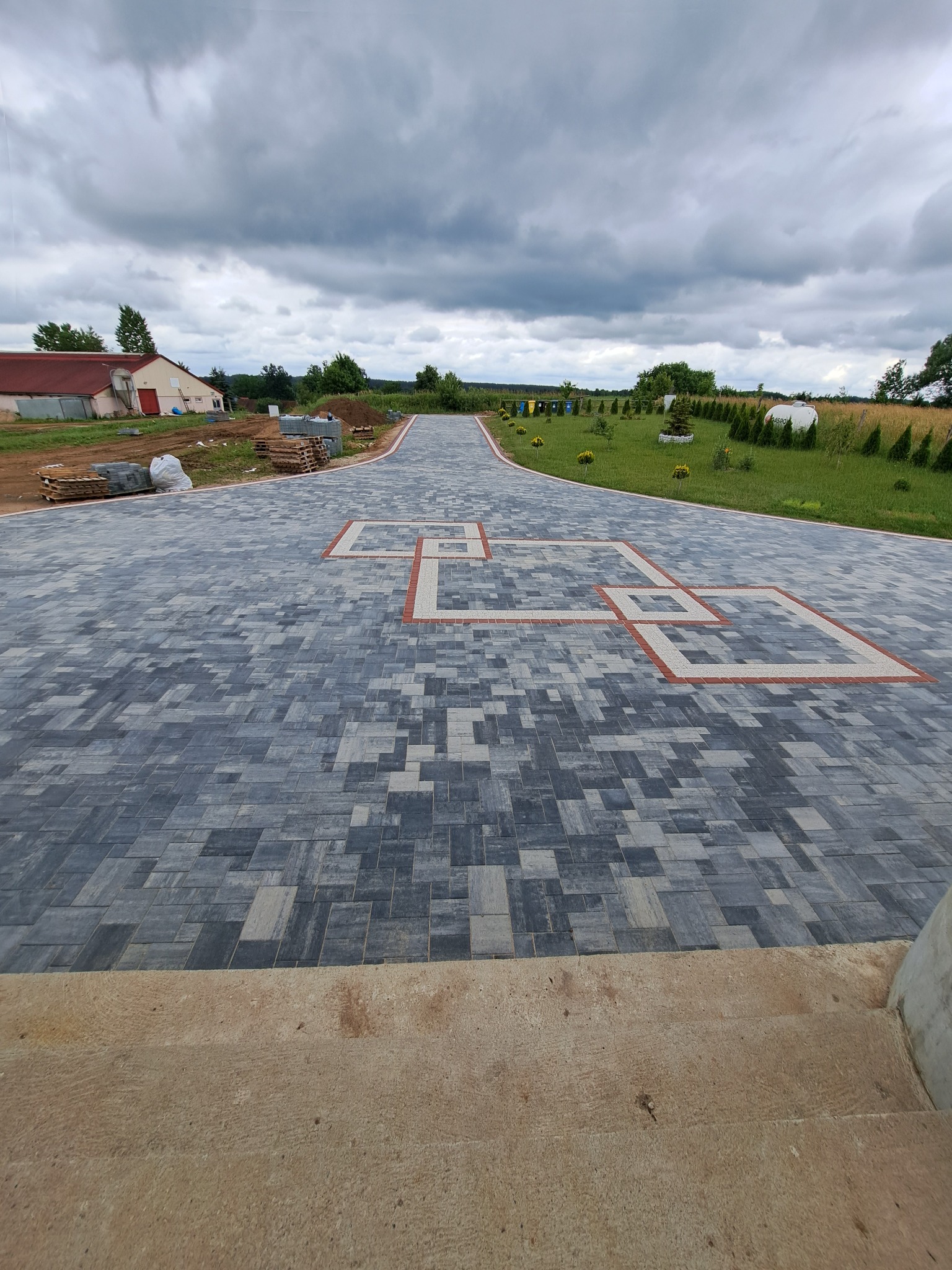 Szeroki podjazd z szarej kostki brukowej ułożonej we wzór, z dekoracyjnymi elementami w kolorze czerwonym i białym. W tle budynek i zielony trawnik pod pochmurnym niebem.