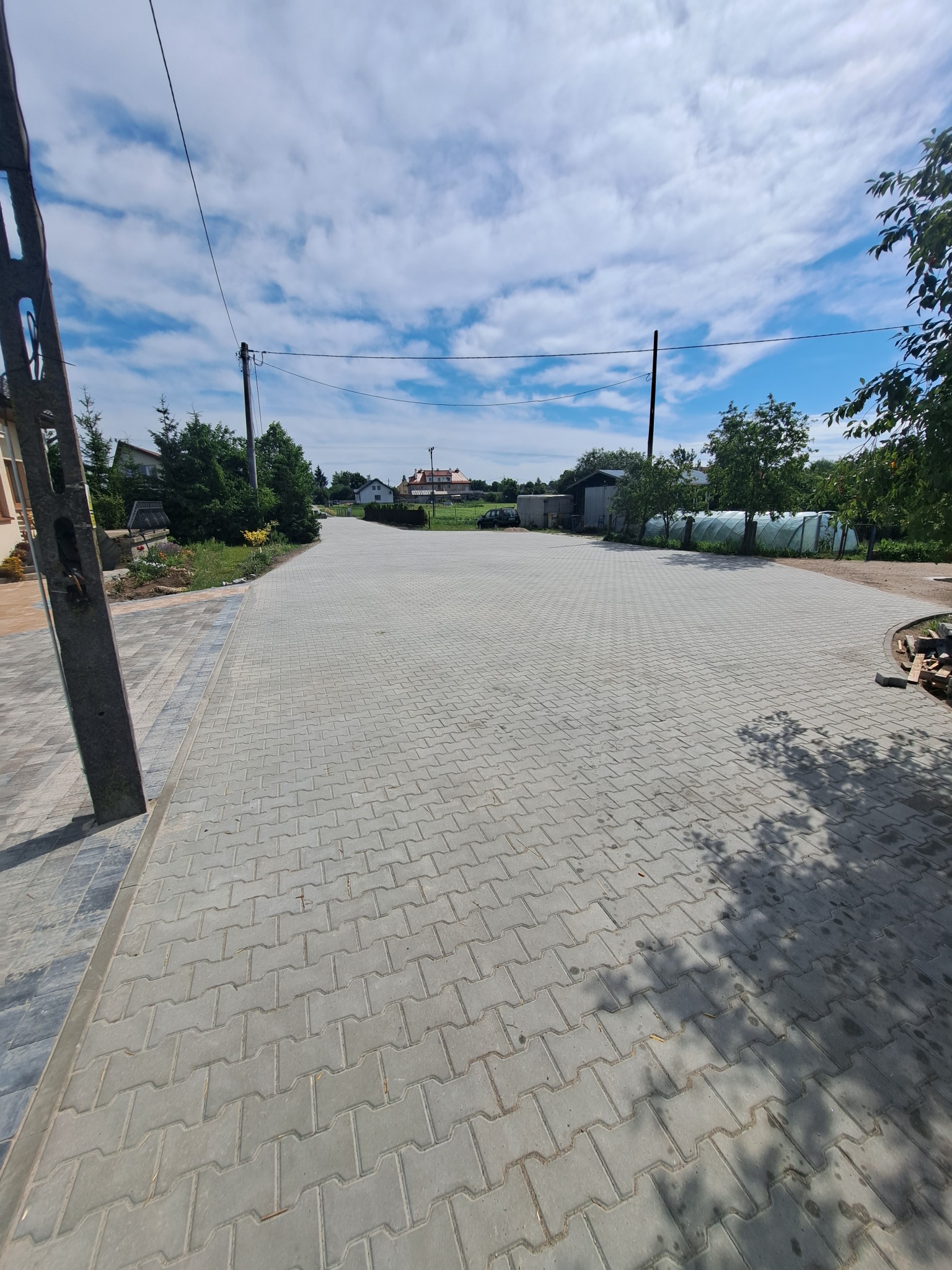 Szeroki podjazd z szarej kostki brukowej w kształcie litery H, prowadzący do domów w oddali, z widocznymi słupami energetycznymi i zielenią po bokach.