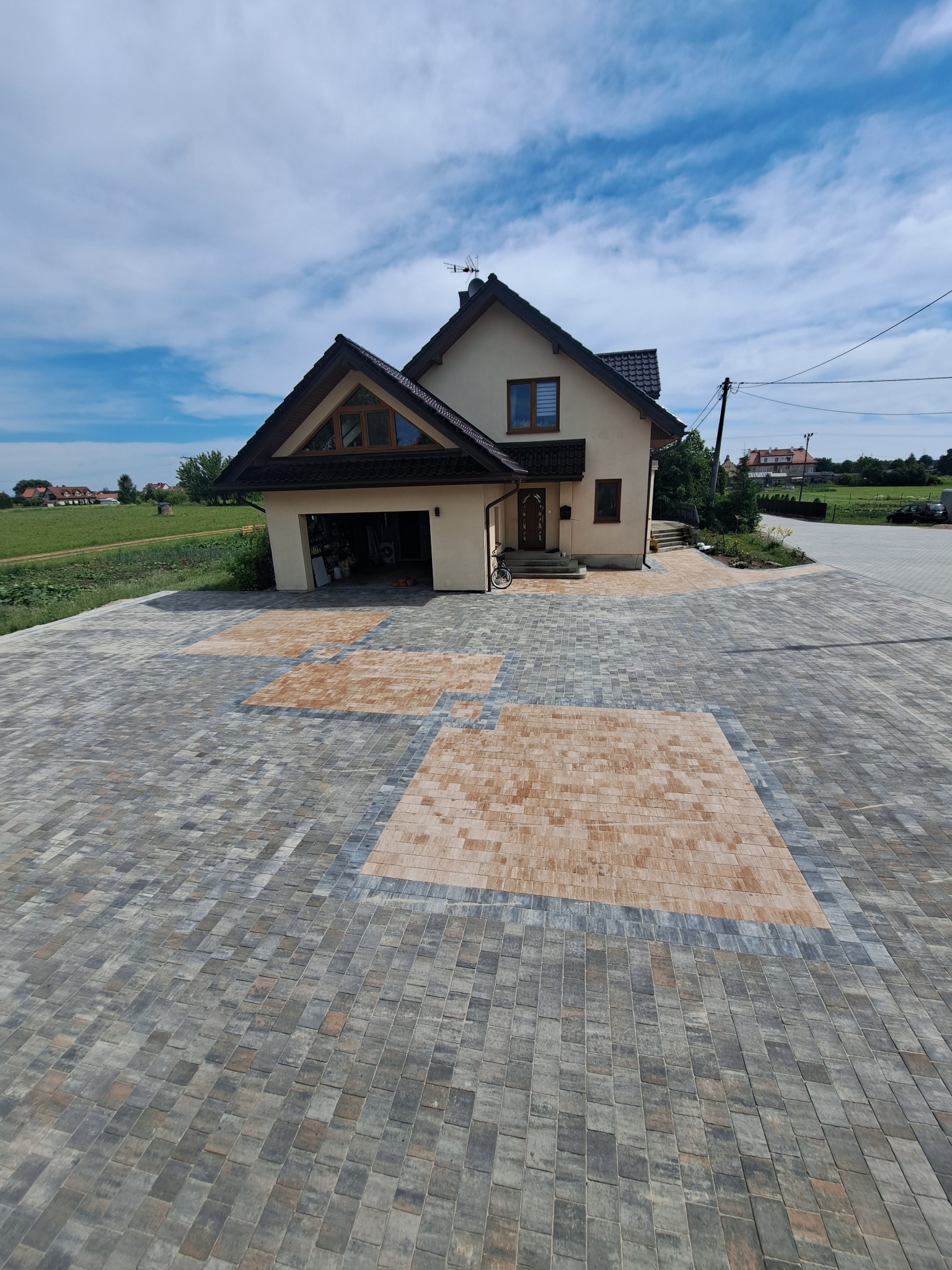 Dom jednorodzinny z garażem i podjazdem z kostki brukowej w odcieniach szarości i beżu, ułożonej we wzór kwadratów. Widok z góry, w tle zielone pola i błękitne niebo.