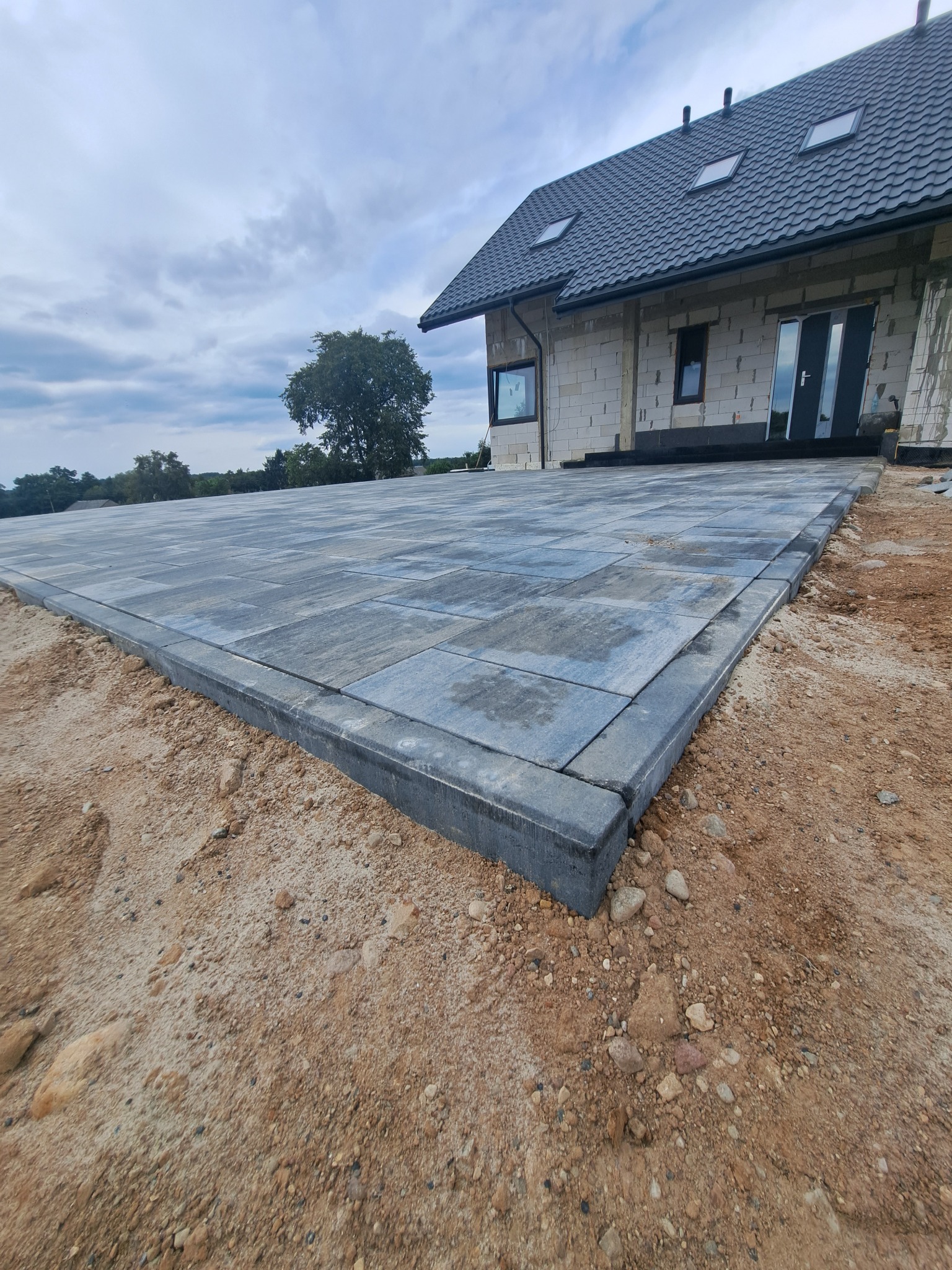 Ułożona kostka brukowa w odcieniach szarości, tworząca taras przed domem w trakcie budowy. Widoczne obrzeża i przygotowane podłoże.