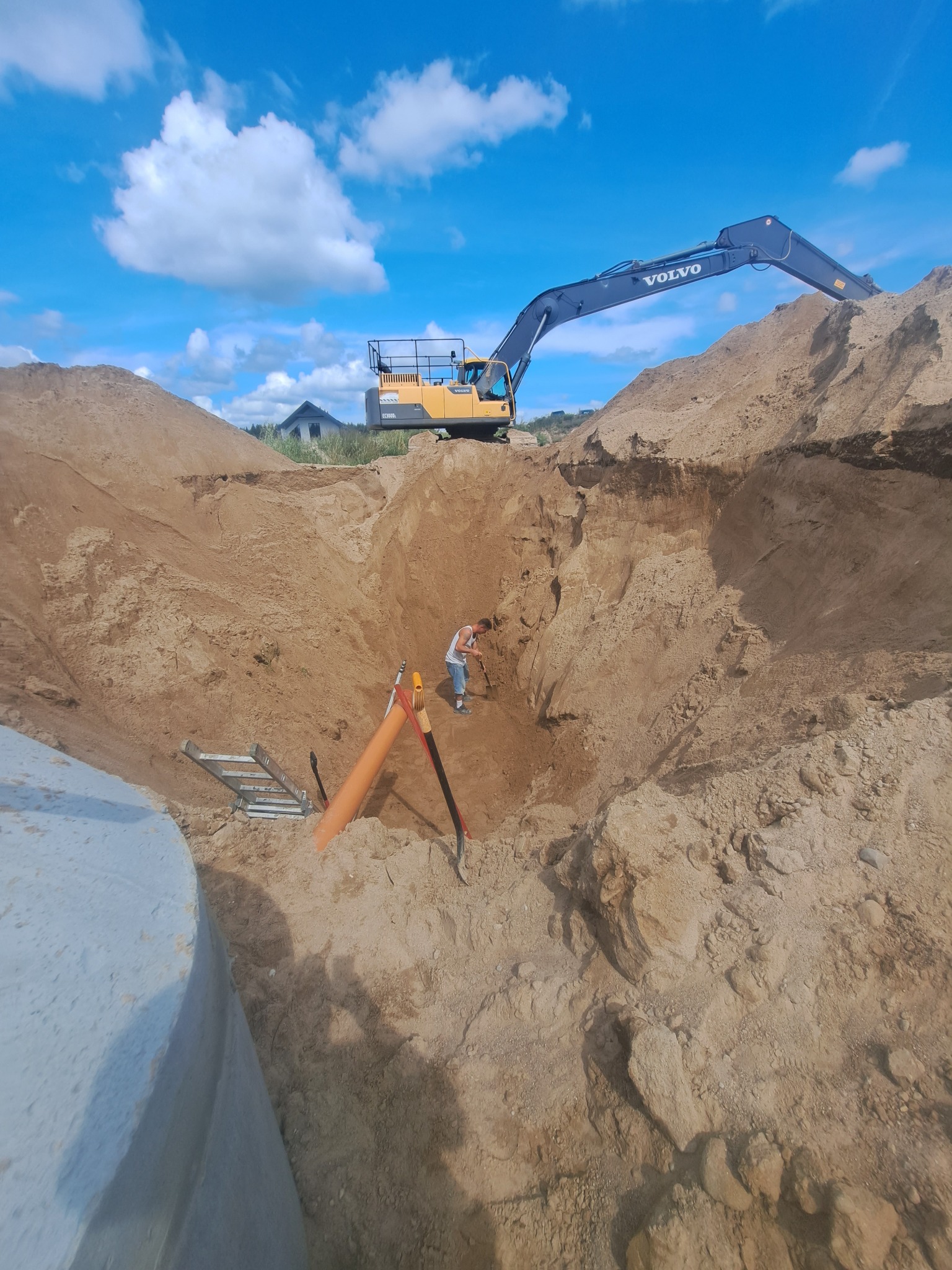 Wykop pod szambo betonowe z widocznym operatorem koparki Volvo na tle błękitnego nieba. W wykopie drabina, rura i narzędzia. Częściowo widoczny betonowy element szamba.