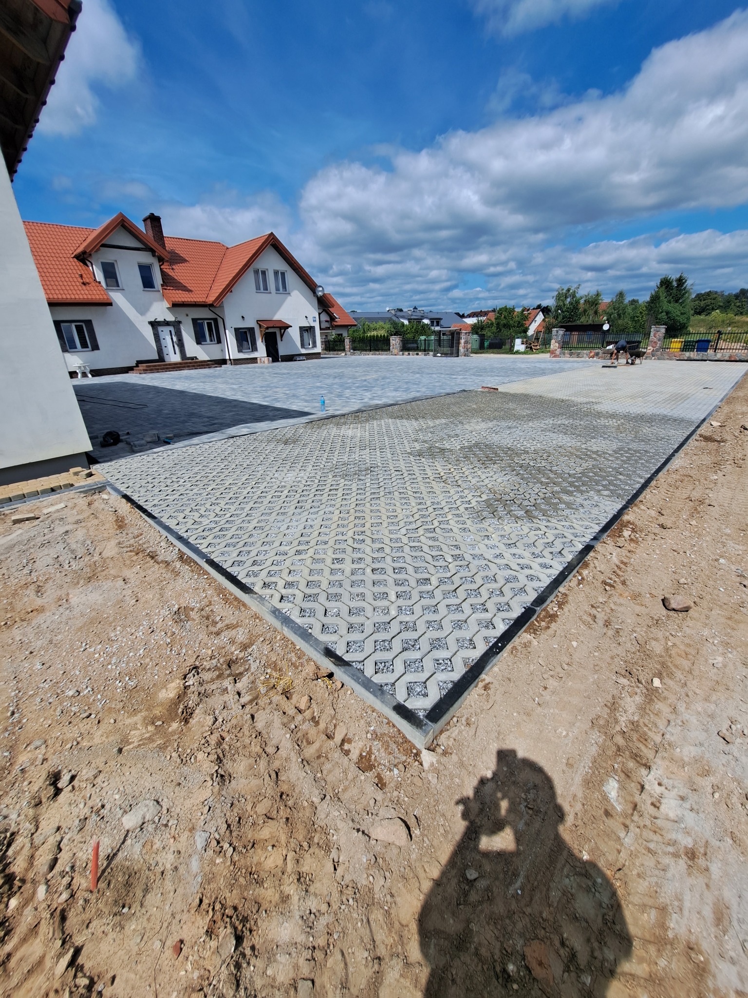 Układanie ażurowej kostki brukowej na posesji z domem w tle. Widoczny fragment ułożonej nawierzchni i przygotowane podłoże. Cień osoby robiącej zdjęcie.