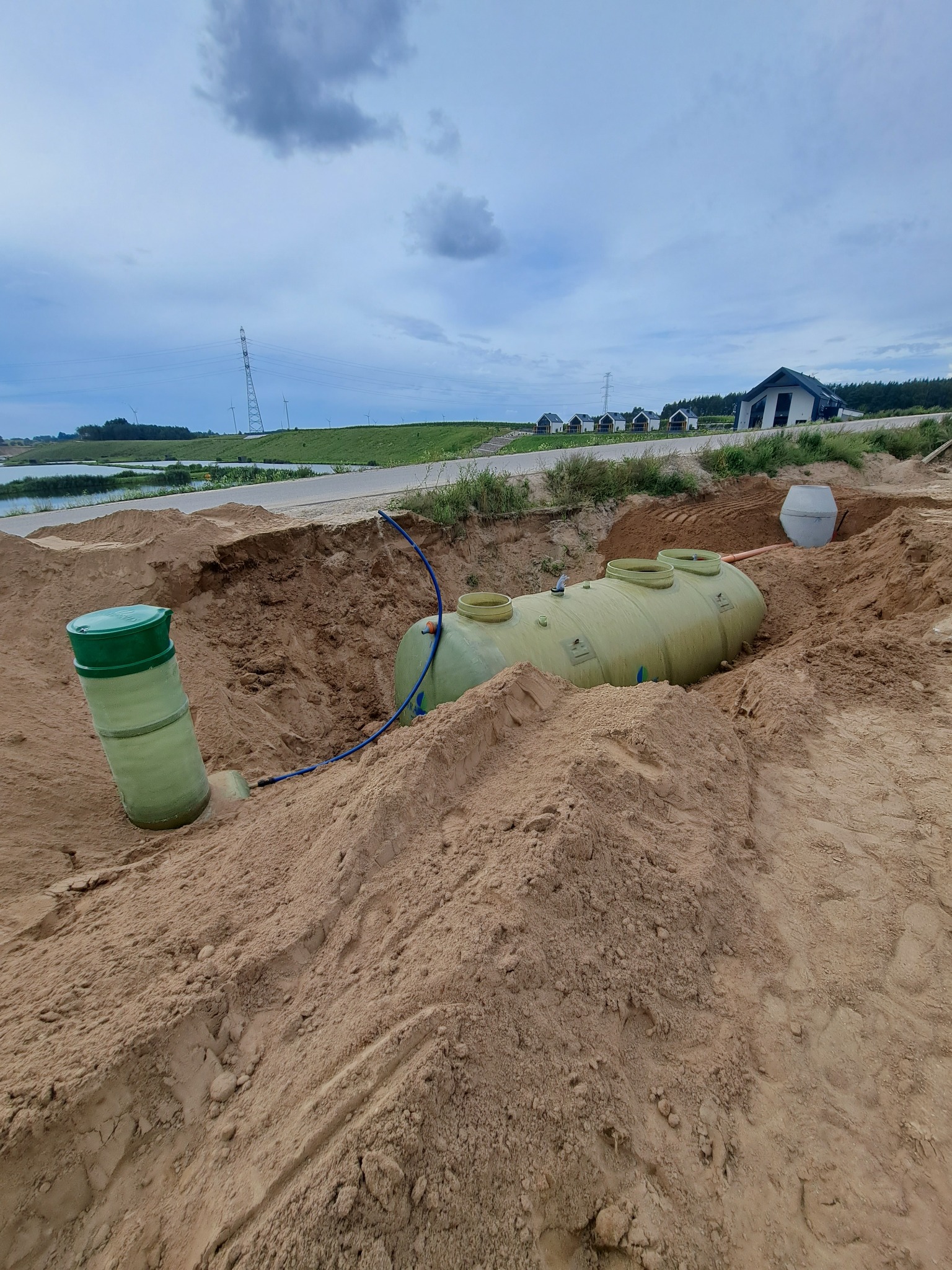 Montaż zbiornika na szambo betonowe w wykopie, widoczne elementy instalacji i krajobraz z domami w tle, pochmurne niebo.