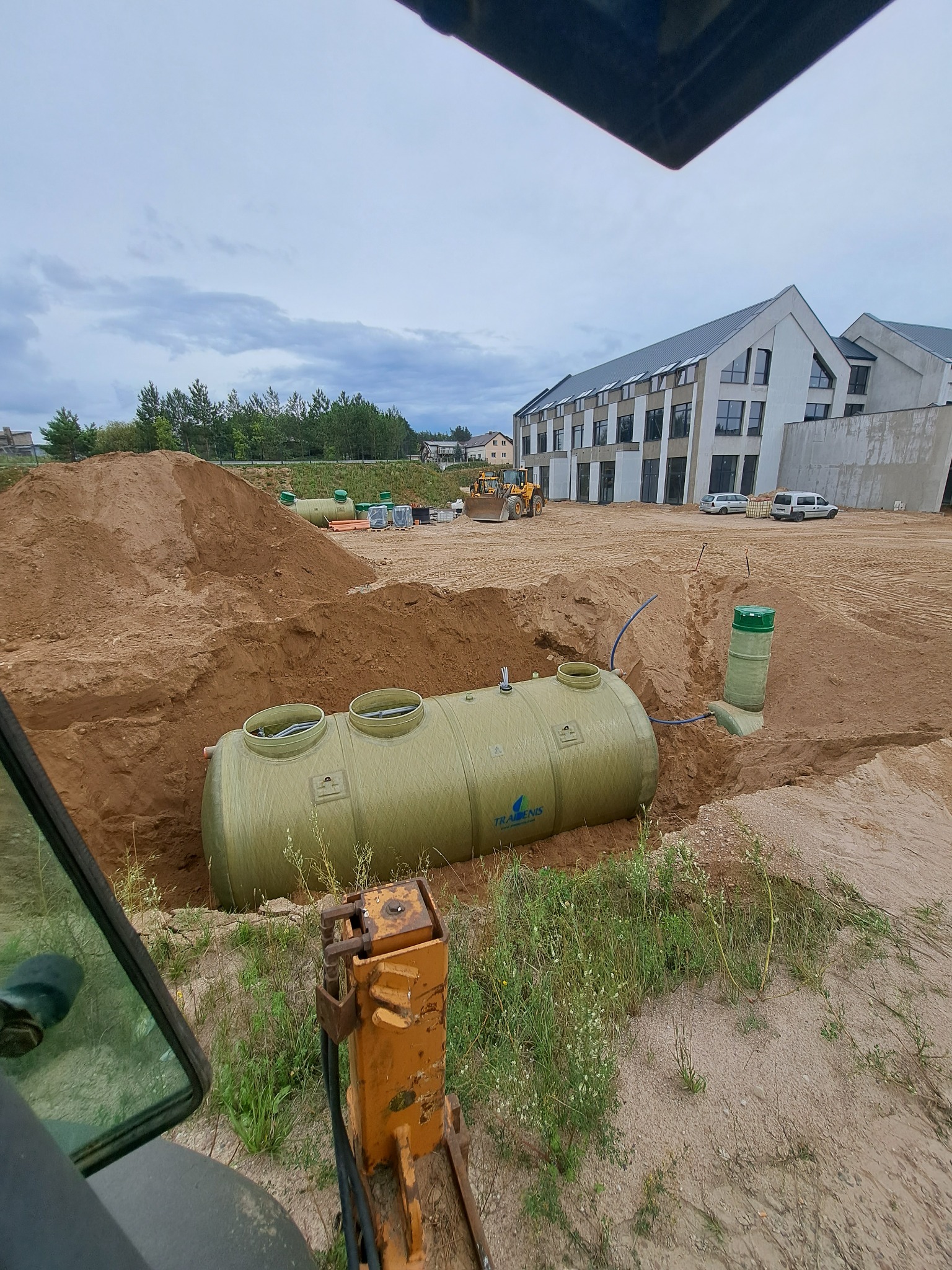 Montaż przydomowej oczyszczalni ścieków w wykopie, widok z kabiny maszyny. W tle budynek i sprzęt budowlany.