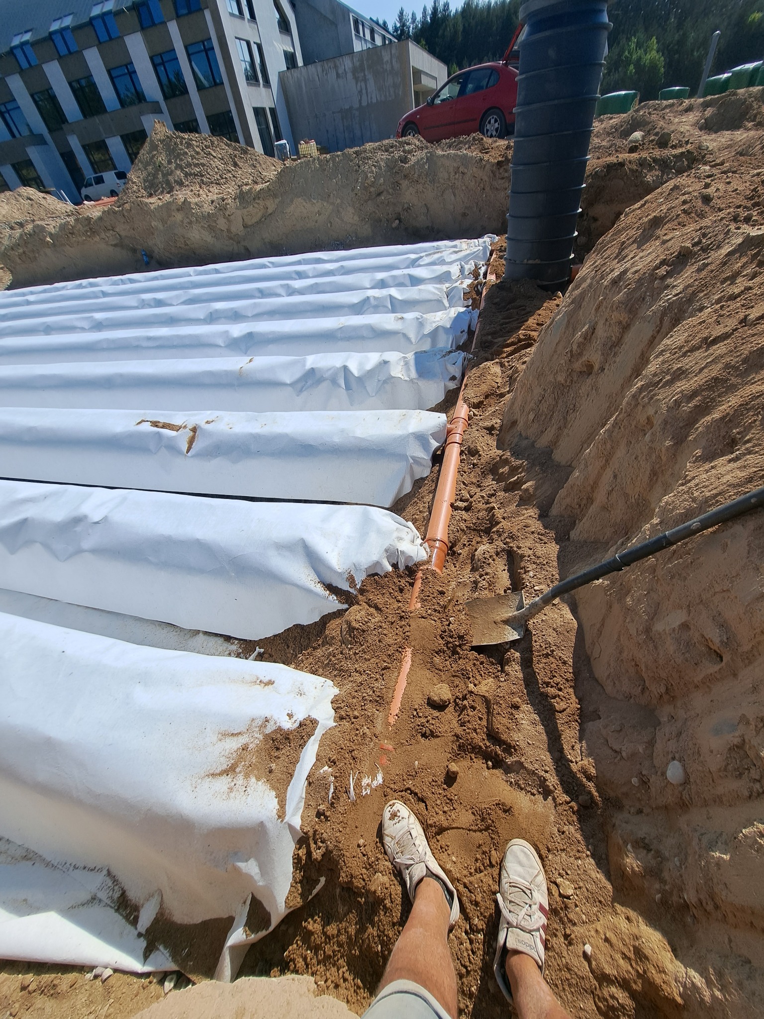 Instalacja drenażu rozsączającego z rur owiniętych geowłókniną w wykopie, widok z góry z perspektywy osoby stojącej w wykopie, z widoczną łopatą i fragmentem budynku w tle.