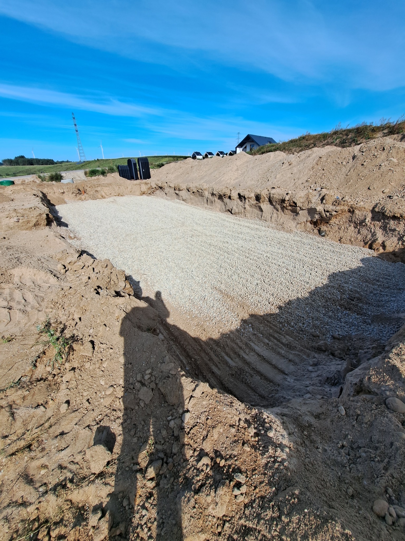 Wykonany wykop pod instalację z podsypką żwirową, widoczne czarne rury i cień osoby. W tle domy i turbiny wiatrowe pod błękitnym niebem.