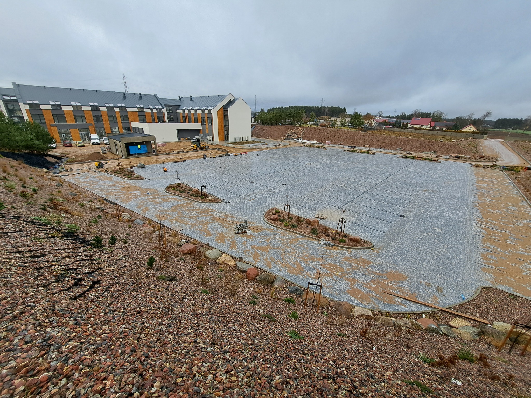 Rozległy parking z szarej kostki brukowej w trakcie układania, z widocznym budynkiem w tle i elementami krajobrazu, takimi jak kamienie i nasadzenia.